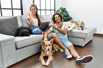 Young hispanic couple with dogs relaxing at home with a big smile on face, pointing with hand finger to the side looking at the camera.