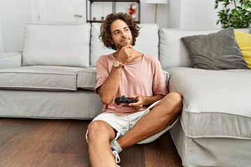 Obraz premium Young hispanic man playing video game holding controller at home serious face thinking about question with hand on chin, thoughtful about confusing idea