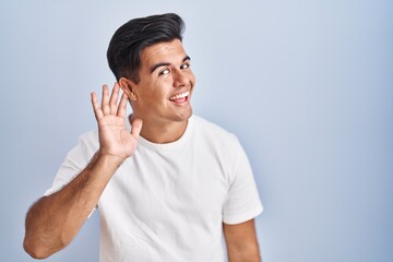 Hispanic man standing over blue background smiling with hand over ear listening an hearing to rumor or gossip. deafness concept.