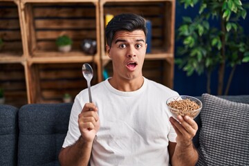 Hispanic man eating healthy whole grain cereals with spoon in shock face, looking skeptical and...