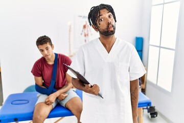 Obraz premium Young hispanic man working at pain recovery clinic with a man with broken arm making fish face with lips, crazy and comical gesture. funny expression.