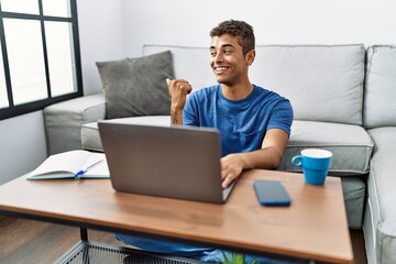 Young handsome hispanic man using laptop sitting on the floor smiling with happy face looking and pointing to the side with thumb up.