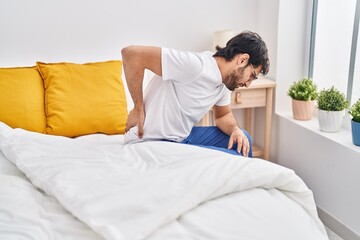 Young hispanic man suffering for back ache sitting on bed at bedroom