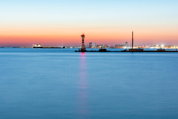 A landscape of sea with a lighthouse in the center and city lights in the foreground. At sunset.