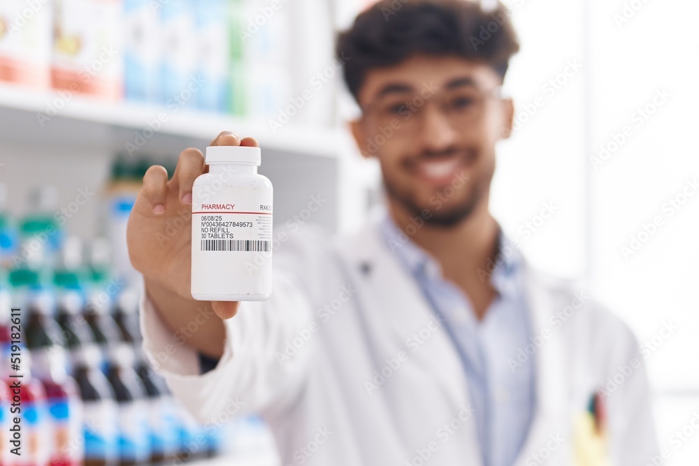 Sticker young arab man pharmacist smiling confident holding pills bottle at pharmacy