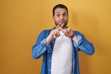 Hispanic man standing over yellow background rejection expression crossing fingers doing negative sign
