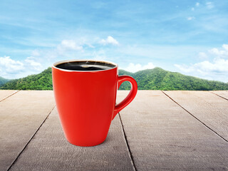 Coffee in a red mug placed on a wooden table with mountains and sky as a beautiful background.