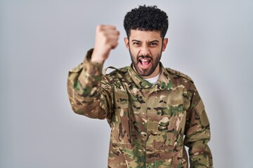 Arab man wearing camouflage army uniform angry and mad raising fist frustrated and furious while...