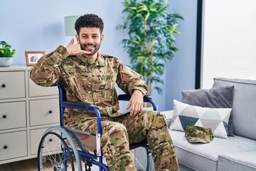 Fototapeta premium Arab man wearing camouflage army uniform sitting on wheelchair smiling doing phone gesture with hand and fingers like talking on the telephone. communicating concepts.