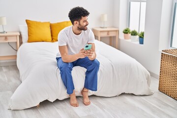 Young arab man using smartphone sitting on bed at bedroom