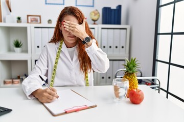 Young redhead woman nutritionist doctor at the clinic covering eyes with hand, looking serious and...