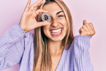 Beautiful hispanic woman holding virtual currency bitcoin screaming proud, celebrating victory and...