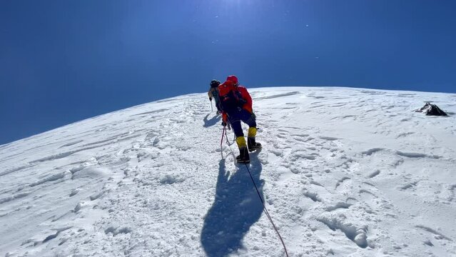 Last steps before Kazbek (Kazbegi) summit 5054m rope team dressed mountaineering clothes, boots with crampons ascending by snowy slope with blue sky background and bright sun. Caucasus mountains. 4K