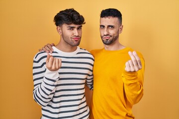 Young hispanic gay couple standing over yellow background doing money gesture with hands, asking...