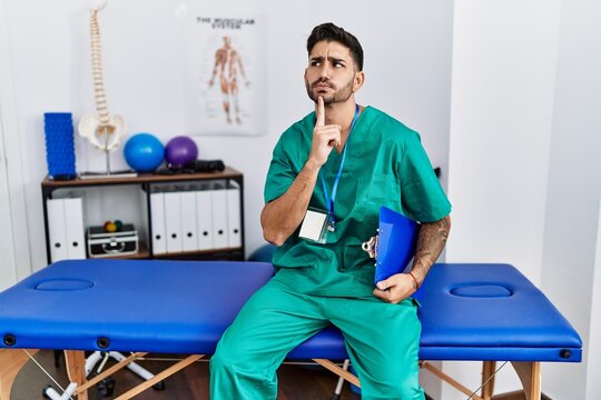 Young physiotherapist man working at pain recovery clinic thinking concentrated about doubt with finger on chin and looking up wondering