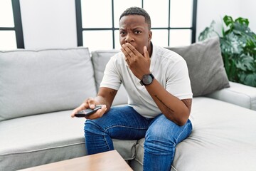 Young african man watching tv holding television remote control covering mouth with hand, shocked...