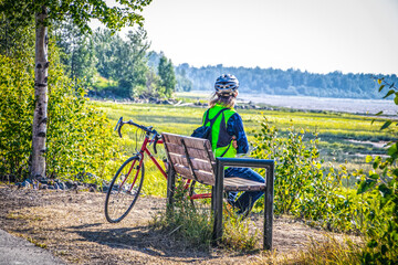 Long haired person in blue jeans with backpack and helment sits looking out at bay from park bench near downtown Anchorage Alaska in summertime - Powered by Adobe