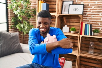 Young african american man hugging piggy bank sitting on sofa at home