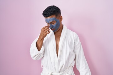 Young hispanic man wearing beauty face mask and bath robe tired rubbing nose and eyes feeling fatigue and headache. stress and frustration concept.