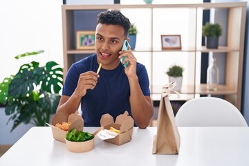 Young latin man talking on smartphone eating take away food at home