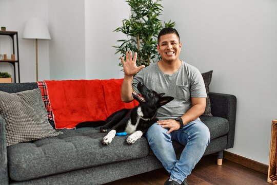 Young latin man and dog sitting on the sofa at home showing and pointing up with fingers number five while smiling confident and happy.