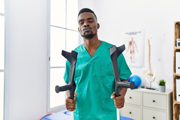 Young african physiotherapist man holding crutches looking at the camera blowing a kiss being...