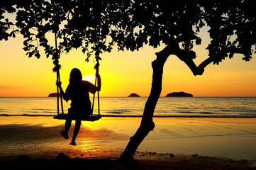 Back view backlight silhouette of a girl swinging at sunset on the beach, THAILAND.