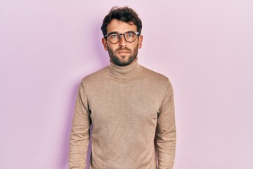 Handsome man with beard wearing turtleneck sweater and glasses relaxed with serious expression on face. simple and natural looking at the camera.