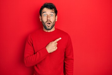 Handsome man with beard wearing casual red sweater surprised pointing with finger to the side, open...