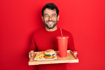 Handsome man with beard eating a tasty classic burger with fries and soda smiling with a happy and...