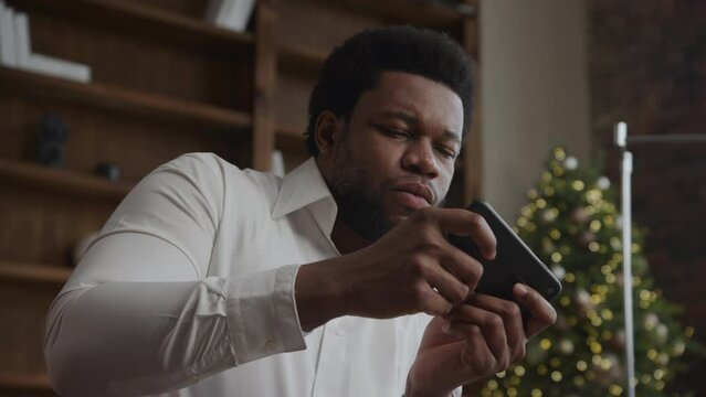 Close-up portrait African-American man playing on smart phone online with Christmass Tree background