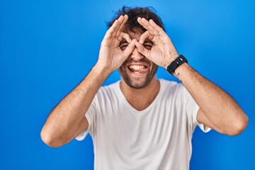 Hispanic young man standing over blue background doing ok gesture like binoculars sticking tongue out, eyes looking through fingers. crazy expression.