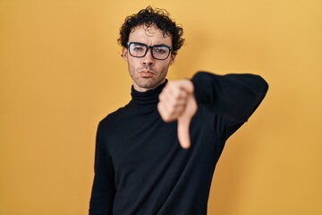 Hispanic man standing over yellow background looking unhappy and angry showing rejection and negative with thumbs down gesture. bad expression.