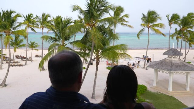 Husband and wife enjoying beach scenery from balcony. Vacationing at Caribbean resort. On third floor overlooking white sand and ocean.