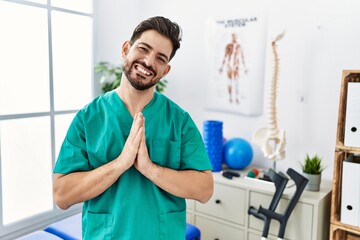 Young man with beard working at pain recovery clinic praying with hands together asking for forgiveness smiling confident.
