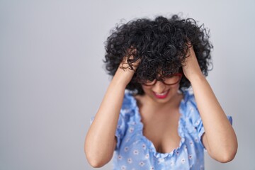 Young brunette woman with curly hair wearing glasses over isolated background suffering from headache desperate and stressed because pain and migraine. hands on head.