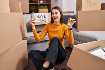 Young caucasian woman sitting on the floor at new home holding new home banner winking looking at...