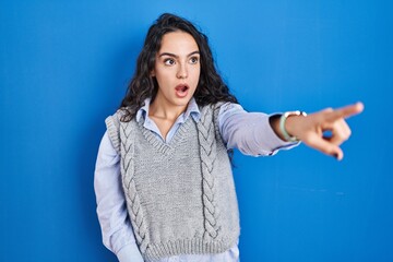 Young brunette woman standing over blue background pointing with finger surprised ahead, open mouth amazed expression, something on the front