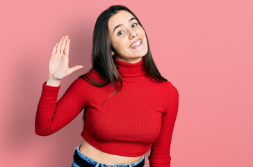 Young brunette teenager wearing red turtleneck sweater waiving saying hello happy and smiling, friendly welcome gesture