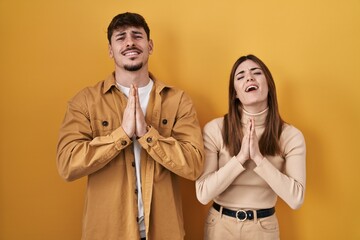 Young hispanic couple standing over yellow background begging and praying with hands together with hope expression on face very emotional and worried. begging.