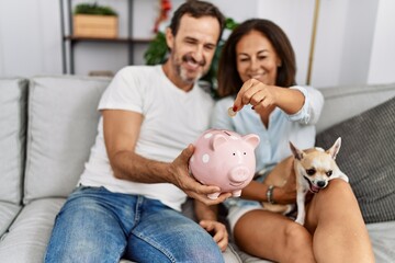 Middle age man and woman couple holding piggy bank sitting on sofa with dog at home
