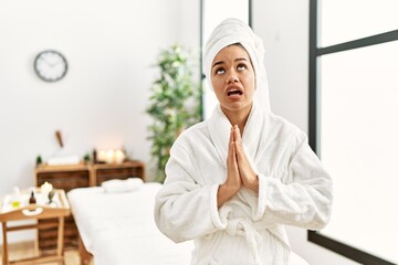 Young brunette woman wearing towel and bathrobe standing at beauty center begging and praying with hands together with hope expression on face very emotional and worried. begging.