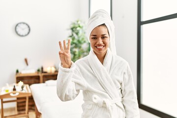 Young brunette woman wearing towel and bathrobe standing at beauty center showing and pointing up with fingers number three while smiling confident and happy.