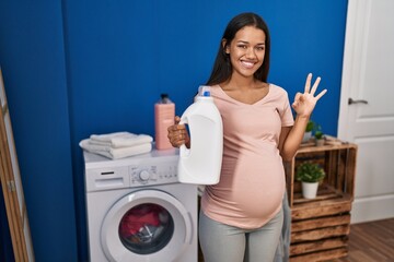 Young pregnant woman doing laundry holding detergent bottle doing ok sign with fingers, smiling friendly gesturing excellent symbol