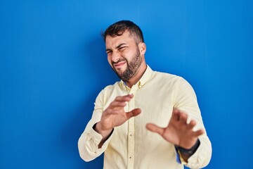 Handsome hispanic man standing over blue background disgusted expression, displeased and fearful...