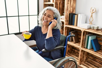 Middle age grey-haired disabled woman talking on the smartphone and drinking coffee sitting on...