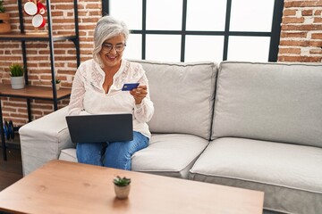 Middle age woman using laptop and credit card sitting on sofa at home
