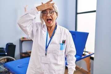 Middle age woman with grey hair working at pain recovery clinic surprised with hand on head for...