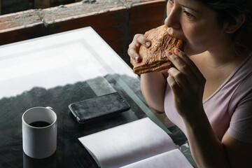 beautiful latina woman eating a sandwich for breakfast before going to college, drinking coffee in...