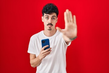 Hispanic man using smartphone over red background doing stop sing with palm of the hand. warning...